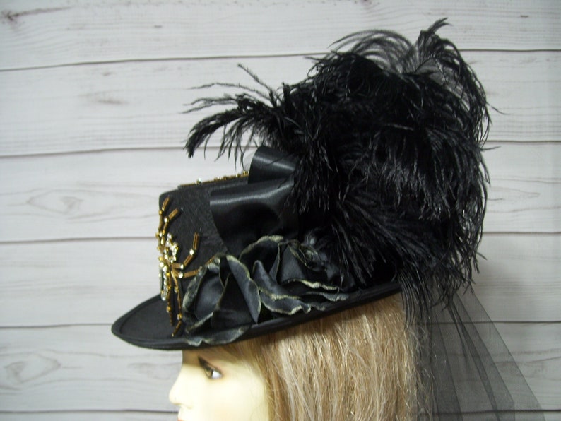 Puede incluir: Un sombrero de copa negro con un dise&ntilde;o de ara&ntilde;a dorado y negro, plumas negras y un velo negro.