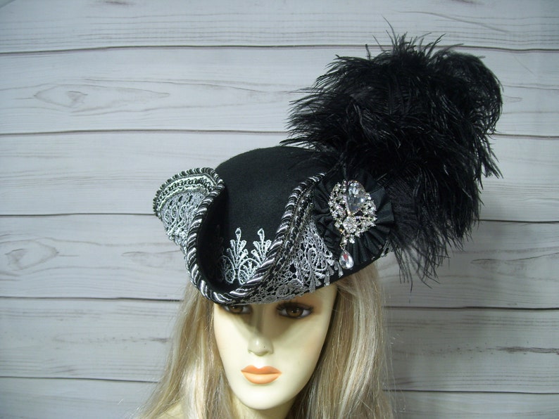 May include: A black felt hat with a silver lace trim and a black feather. The hat has a large, sparkling rhinestone brooch.