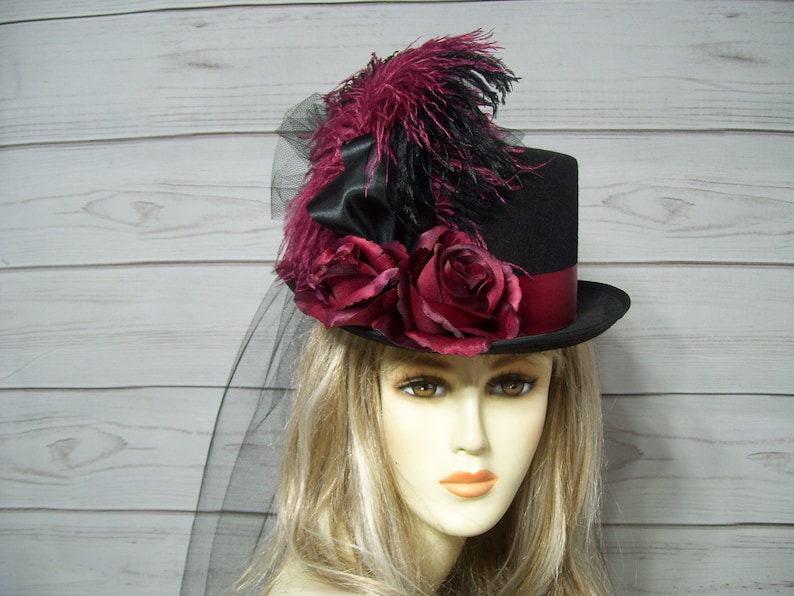 May include: A black top hat with a burgundy ribbon and a veil. The hat is decorated with burgundy roses and black feathers.