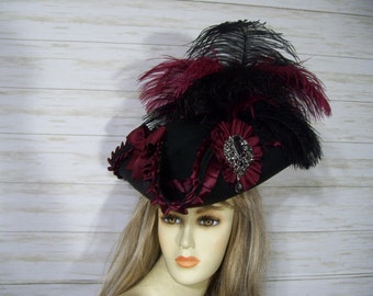 May include: A black tricorn hat with a burgundy ribbon and a large jeweled brooch. The hat is adorned with black and burgundy feathers.