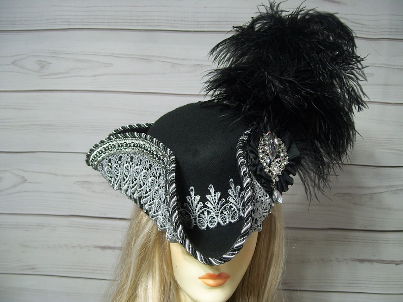 May include: A black felt tricorn hat with a silver lace trim and a black feather plume. The hat has a jeweled embellishment on the side.