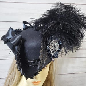 Pu&ograve; includere: Un cappello nero con una corona quadrata, adornato con un grande gruppo di piume nere. Il cappello presenta un nastro decorativo, finiture a balze e un accento di gioiello scintillante. Il cappello &egrave; progettato con un'estetica vintage.