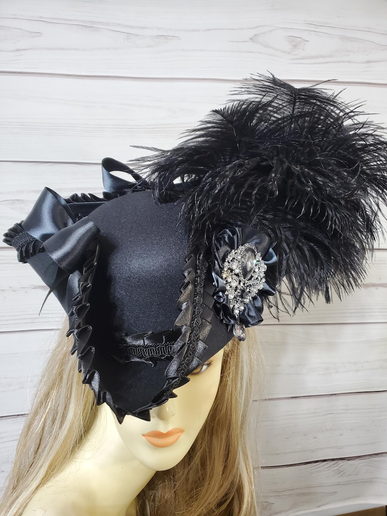 Pu&ograve; includere: Un cappello nero con una corona quadrata, adornato con un grande gruppo di piume nere. Il cappello presenta un nastro decorativo, finiture a balze e un accento di gioiello scintillante. Il cappello &egrave; progettato con un'estetica vintage.