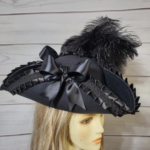 Pu&ograve; includere: Un cappello nero con tesa larga, ornato da un grande fiocco di raso e piume nere. Il bordo &egrave; rifinito con un bordo nero decorativo. Il cappello &egrave; di colore scuro e ha uno stile vintage o teatrale.