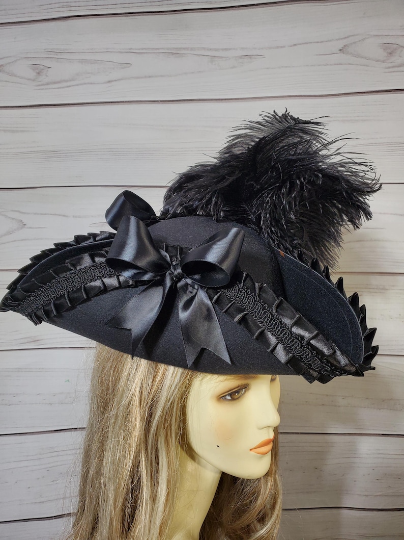 Pu&ograve; includere: Un cappello nero con tesa larga, ornato da un grande fiocco di raso e piume nere. Il bordo &egrave; rifinito con un bordo nero decorativo. Il cappello &egrave; di colore scuro e ha uno stile vintage o teatrale.