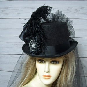 May include: A black top hat with a black feather, black satin ribbon, and a rhinestone embellishment. The hat is adorned with black tulle and a black veil.