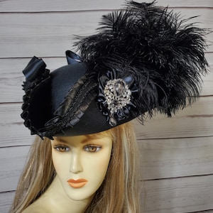 Pu&ograve; includere: Un cappello nero con tesa larga, ornato da un grande gruppo di piume nere. Il cappello presenta un nastro decorativo, un abbellimento gioiello e un bordo a balze. Il cappello &egrave; esposto su un manichino con capelli biondi.