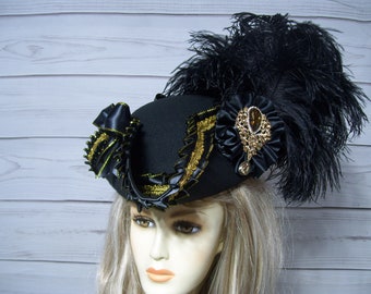 May include: A black felt hat with a wide brim, gold trim, and a black feather plume. The hat is adorned with a black satin bow and a jeweled embellishment.