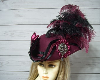 May include: A burgundy felt hat with a wide brim and a black and burgundy feather plume. The hat is decorated with black ribbon and a jeweled brooch.