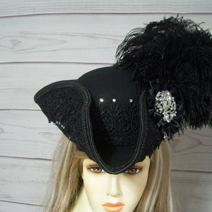 Puede incluir: Un sombrero de fieltro negro con ala ancha y una pluma negra. El sombrero está decorado con encaje negro y un gran broche de pedrería.