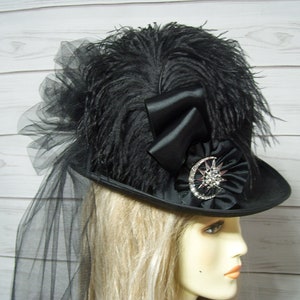 May include: A black hat with a large black feather plume, a black satin bow, and a black and silver starburst embellishment. The hat is worn by a person with blonde hair.