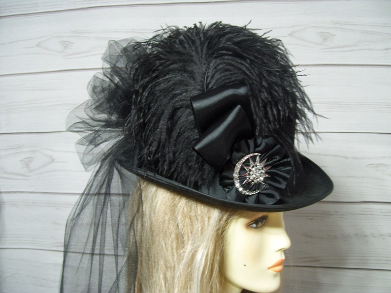 May include: A black hat with a large black feather plume, a black satin bow, and a black and silver starburst embellishment. The hat is worn by a person with blonde hair.