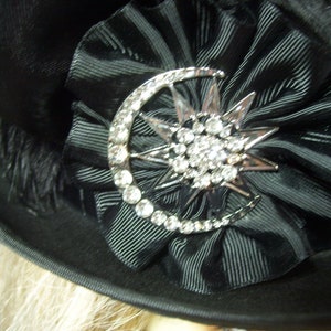May include: A black hat with a black satin ribbon and a silver crescent moon and star brooch with rhinestones.