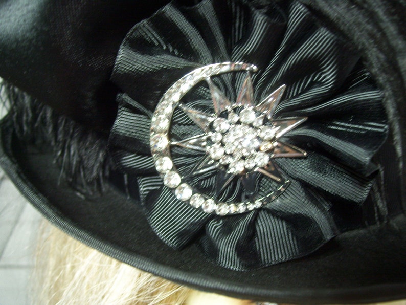May include: A black hat with a black satin ribbon and a silver crescent moon and star brooch with rhinestones.