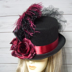 May include: A black top hat with a red ribbon and a large burgundy rose on the side. The hat is decorated with black and burgundy feathers and a black tulle veil.