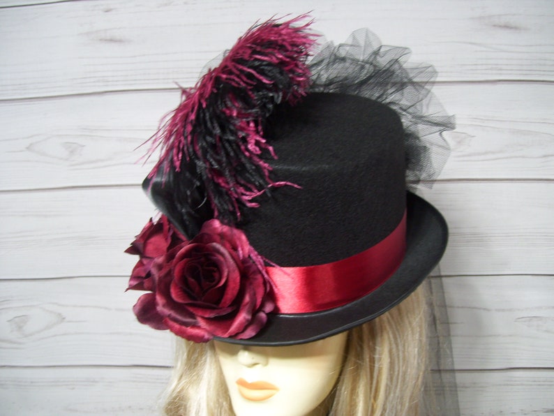 May include: A black top hat with a red ribbon and a large burgundy rose on the side. The hat is decorated with black and burgundy feathers and a black tulle veil.