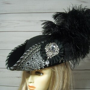 May include: A black felt pirate hat with a silver trim and a large black feather. The hat has a large rhinestone brooch on the front.