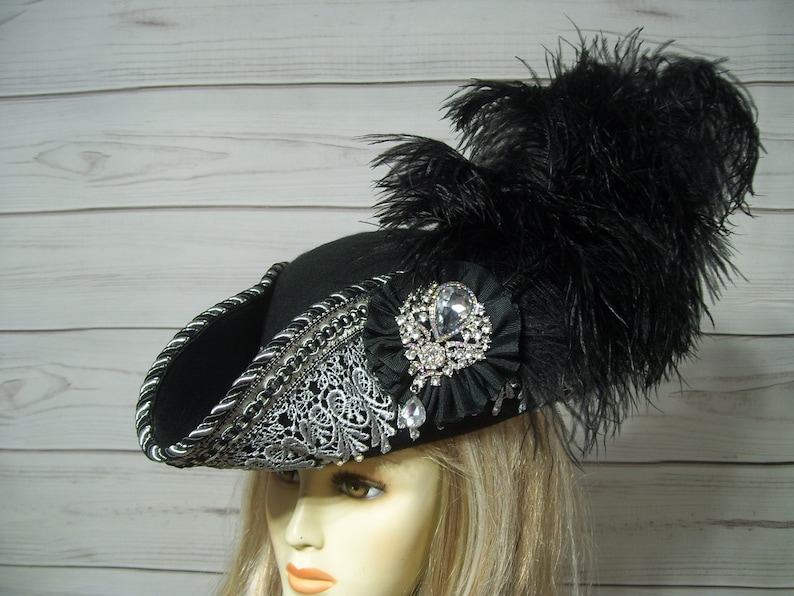 May include: A black felt pirate hat with a silver trim and a large black feather. The hat has a large rhinestone brooch on the front.