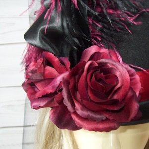 May include: A black top hat with a red ribbon and a cluster of burgundy roses. The hat is decorated with black feathers and a black satin bow.