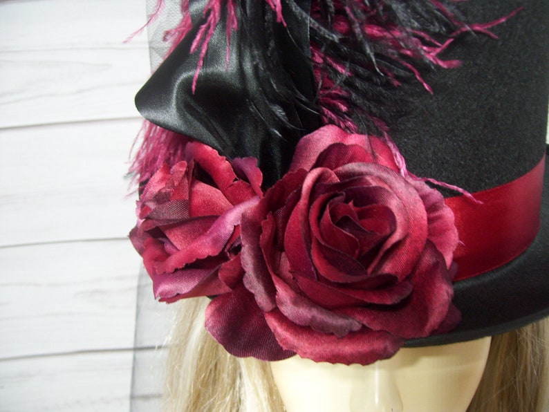 May include: A black top hat with a red ribbon and a cluster of burgundy roses. The hat is decorated with black feathers and a black satin bow.