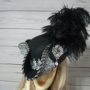 May include: A black felt hat with a silver and white lace trim and a black feather plume. The hat has a large, clear rhinestone brooch.