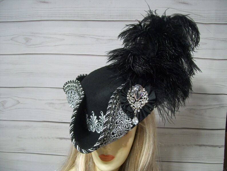 May include: A black felt hat with a silver and white lace trim and a black feather plume. The hat has a large, clear rhinestone brooch.