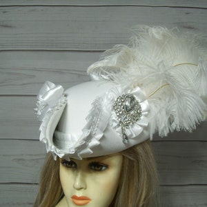 May include: A white hat with a large white feather and a rhinestone embellishment. The hat has a wide brim and is trimmed with white satin ribbon.