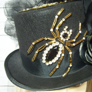 Puede incluir: Un sombrero de copa negro con una banda de sat&eacute;n negro y una rosa negra en el lateral. El sombrero est&aacute; decorado con una ara&ntilde;a grande y enjoyada hecha de cuentas doradas y diamantes de imitaci&oacute;n.