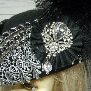 May include: A black felt pirate hat with silver trim and a large black bow adorned with a sparkling rhinestone brooch. The hat is decorated with a silver lace trim and a black feather plume.