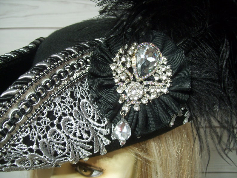 May include: A black felt pirate hat with silver trim and a large black bow adorned with a sparkling rhinestone brooch. The hat is decorated with a silver lace trim and a black feather plume.