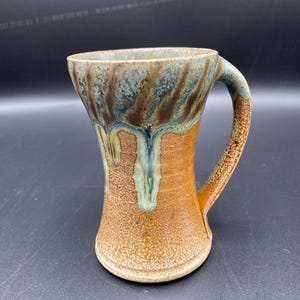 May include: A brown ceramic mug with a blue and green glaze. The mug has a wide base and a narrow top, with a handle on one side.