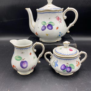 Puede incluir: Un juego de té de porcelana blanca con un diseño floral y de frutas. El juego incluye una tetera, una jarra de crema y un azucarero. La tetera tiene un borde dorado y una tapa con un pomo. La jarra de crema y el azucarero tienen un borde dorado y un diseño floral.