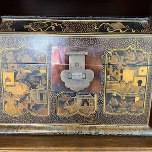 May include: A rectangular black lacquer box featuring intricate gold painted landscape scenes with pagodas and figures. The box has a hinged lid, a central metal latch plate, and a decorative border pattern along the base and top edges.