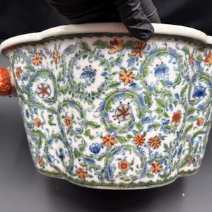 May include: A ceramic bowl with a scalloped rim and a white base featuring a repeating pattern of blue and green scrolling vines, orange flowers, and small blue blossoms. The bowl has two handles shaped like textured red strawberries on each side.