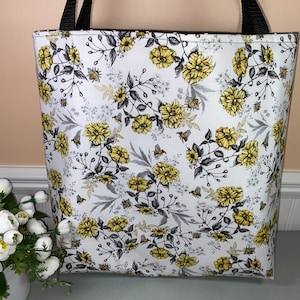 May include: A white tote bag with black straps and a floral print featuring yellow flowers, gray leaves, and small bees.
