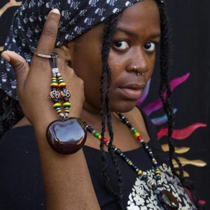 Puede incluir: Una mujer con largas trenzas negras lleva una bandana negra con un patrón de calavera y huesos cruzados. Lleva una camisa negra con un collar de cuentas con un gran colgante de semilla marrón.