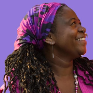 May include: A woman with long, dark brown dreadlocks wears a pink and purple camouflage headband. She is wearing a pink shirt and a beaded necklace with a large, dark brown pendant.