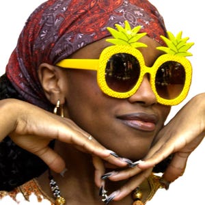 Peut inclure: Une personne porte des lunettes de soleil fantaisie en forme d'ananas jaunes avec des verres bruns. La personne porte également un foulard à motifs rouges et violets et un collier de perles. Les lunettes de soleil sont au centre de l'image.