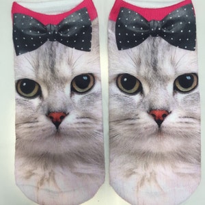 May include: A pair of white ankle socks with a gray cat face and a black bow tie with white polka dots. The bow tie is pink on the top.