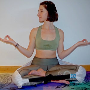 May include: A woman sits in a yoga pose wearing a green sports bra and matching green leggings. She is wearing a white yoga strap around her legs. She is sitting on a colorful yoga mat.