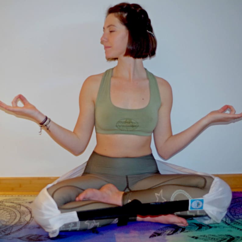 May include: A woman sits in a yoga pose wearing a green sports bra and matching green leggings. She is wearing a white yoga strap around her legs. She is sitting on a colorful yoga mat.
