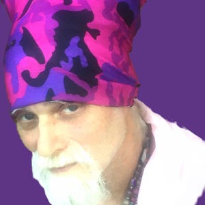 May include: A man with a white beard and a pink, purple, and black camouflage bandana on his head. He is wearing a white shirt with a multi-colored beaded necklace.