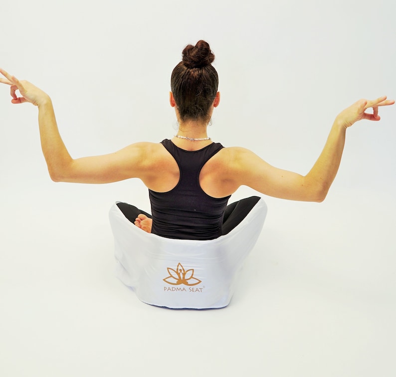 May include: A person sits in a white meditation cushion with a lotus flower logo and the text "PADMA SEAT". The cushion is on a white surface. The person is wearing a black tank top and has their arms raised.