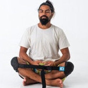May include: A person sits cross-legged on the floor wearing a white t-shirt and black knee pads. A black strap with a buckle is wrapped around their legs.