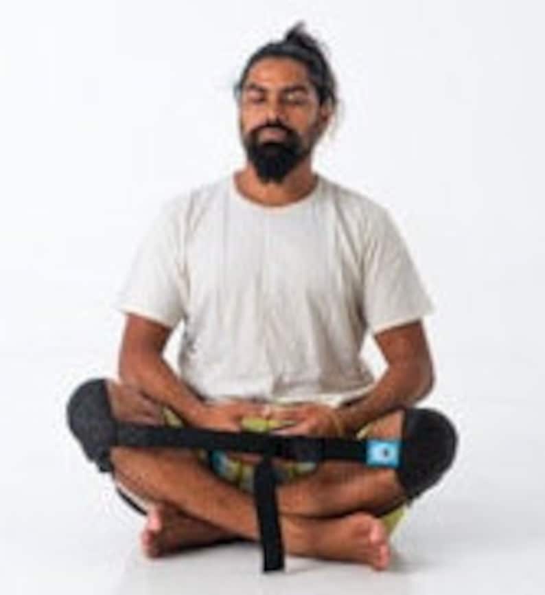 May include: A person sits cross-legged on the floor wearing a white t-shirt and black knee pads. A black strap with a buckle is wrapped around their legs.