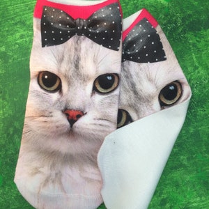 May include: A pair of white ankle socks with a cat face design. The cat is wearing a black and white polka dot bow tie. The socks are on a green background.