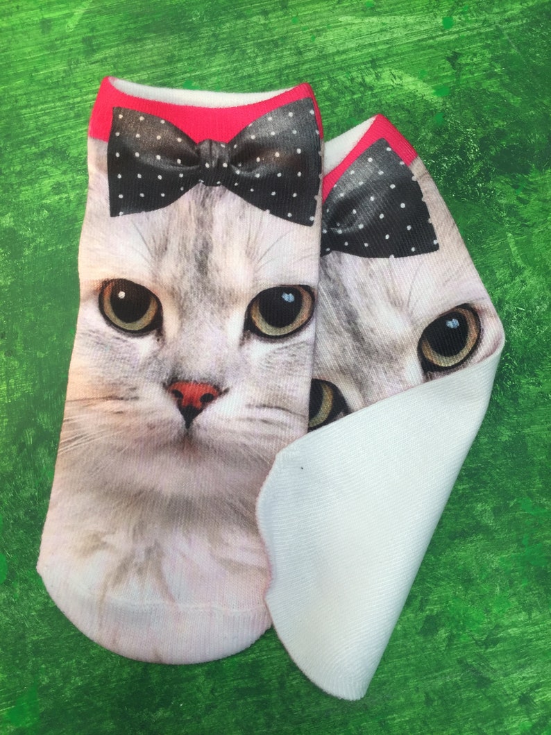 May include: A pair of white ankle socks with a cat face design. The cat is wearing a black and white polka dot bow tie. The socks are on a green background.