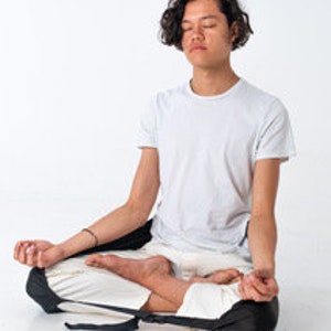 May include: A person sits in a meditative pose wearing black yoga pants with a strap that goes around the knees and white pants underneath. The person is wearing a white t-shirt and has their eyes closed.