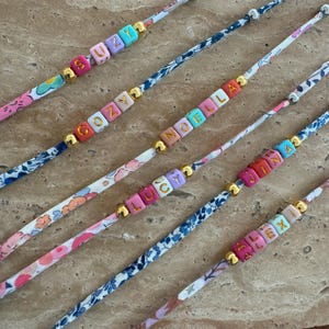 May include: Six colorful bracelets with different names spelled out in gold letters on colorful beads. The bracelets are made with a white fabric with a floral pattern.