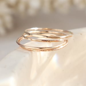 May include: A set of three thin, polished rose gold rings stacked together. Each ring features a smooth, rounded band with a reflective metallic finish. The rings are positioned in a cluster, showcasing their uniform circular shape and warm, pinkish-gold hue.